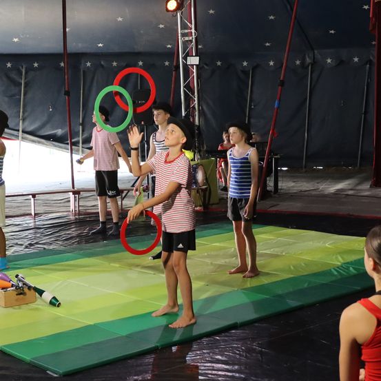 Eine Gruppe junger Kinder übt das Jonglieren bei einer Zirkusveranstaltung. Sie stehen auf einem bunten Teppich unter einem Zelt mit einem Sternenhimmel im Hintergrund.