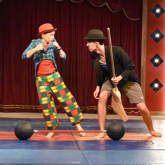Zwei Personen führen eine Darbietung auf der Bühne auf. Eine Person trägt einen Hut und karierte Hosen. Die andere Person trägt einen Hut und hält einen Besen. Sie stehen beide auf einem Balancebalken mit Gewichten an den Enden.