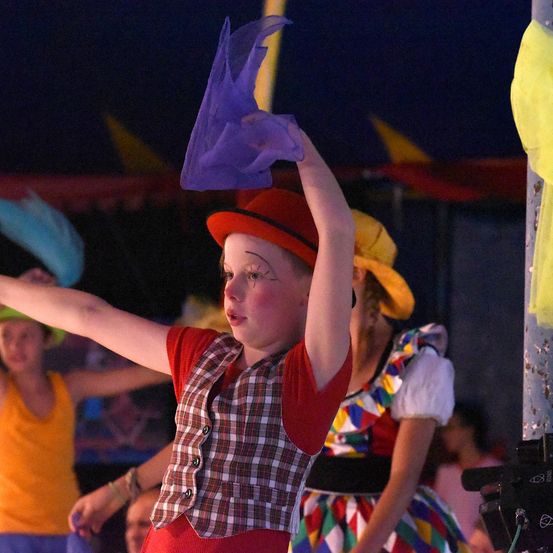 Ein junger Junge in einem rot-weiß karierten Weste und einem roten Hut hält eine lila Flagge während einer Aufführung, mit einer anderen Darstellerin in einem farbenfrohen Kleid und einem gelben Hut hinter ihm.