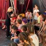 Eine Gruppe von Kindern sitzt auf einer Bank unter einem Zirkuszelt und hört einer Frau zu.