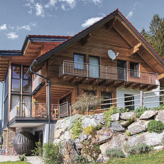Ein Holzhaus mit rotem Dach, zwei Balkonen und einer Satellitenschüssel oben. Es hat Glasfenster und Türen. Es gibt Steine und Pflanzen drumherum. Vorne ist ein Fahrrad geparkt.
