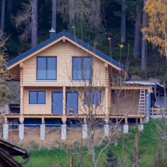 Ein Holzhaus im Bau mit Glasfenstern und einem blauen Dach, auf einem Hang gelegen, umgeben von Bäumen und Pflanzen.