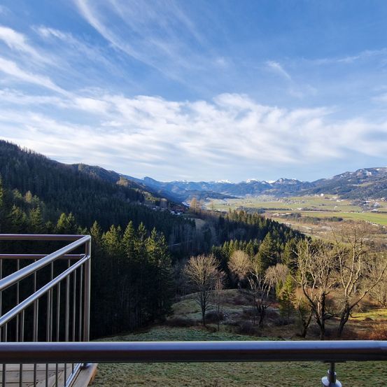 Ein Balkonblick auf ein hügeliges Gebiet mit dichten Bäumen und einer Bergkette in der Ferne. Der Himmel ist blau mit weißen Wolken.