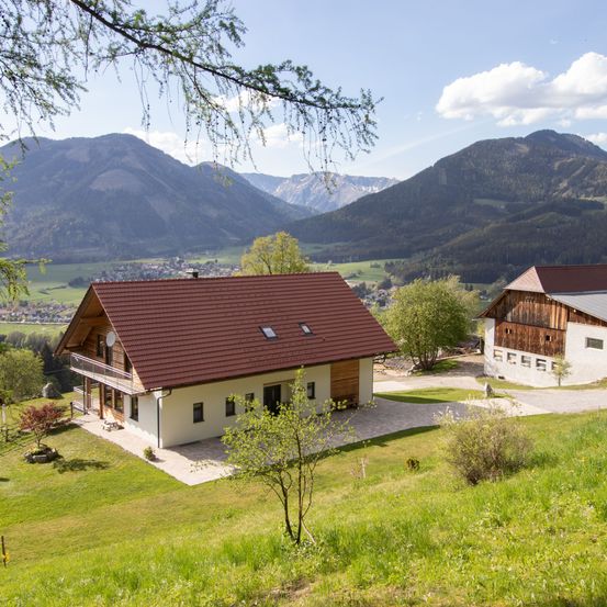 Ein Haus mit einem braunen Dach liegt in einem Tal mit grünen Feldern und Bergen im Hintergrund. Ein weiteres Haus ist rechts sichtbar.