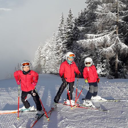 Drei Kinder in roten Jacken und Helmen steigen einen schneebedeckten Hang hinunter. Sie lächeln und scheinen für ein Foto zu posieren.