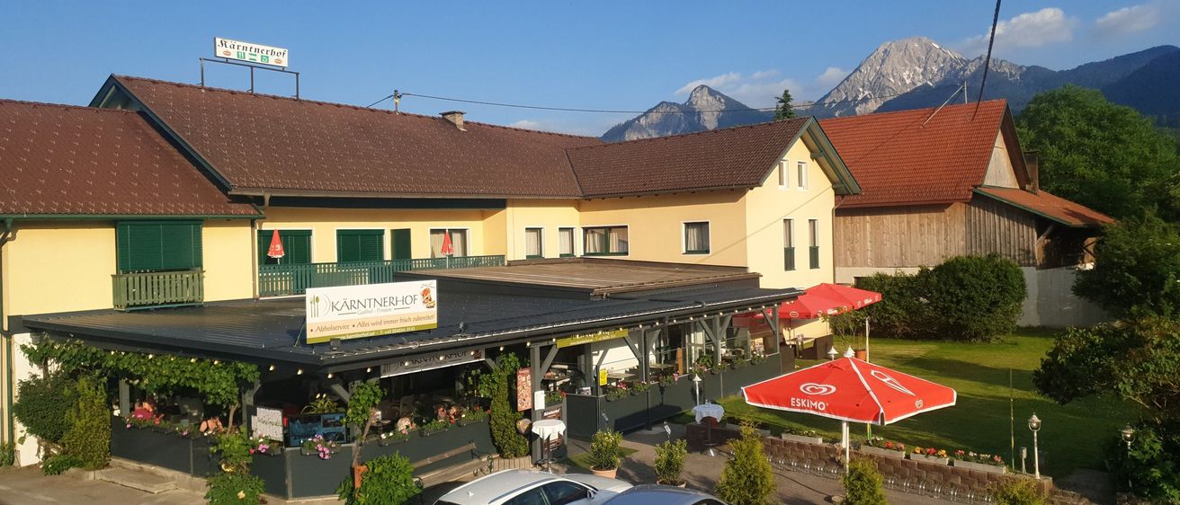 Ein Restaurant mit Sitzgelegenheiten im Freien und Bergen im Hintergrund. Das Restaurant hat eine gelbe Außenfassade und grüne Fensterläden. Das Restaurant-Schild ist blau mit weißen Buchstaben.