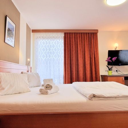 Ein Schlafzimmer mit einem ordentlich gemachten Bett und weißen Kissen. Ein Bilderrahmen ist an der Wand, und ein Blumentopf steht auf einem Schreibtisch.