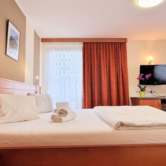 Ein Schlafzimmer mit einem ordentlich gemachten Bett und weißen Kissen. Ein Bilderrahmen ist an der Wand, und ein Blumentopf steht auf einem Schreibtisch.