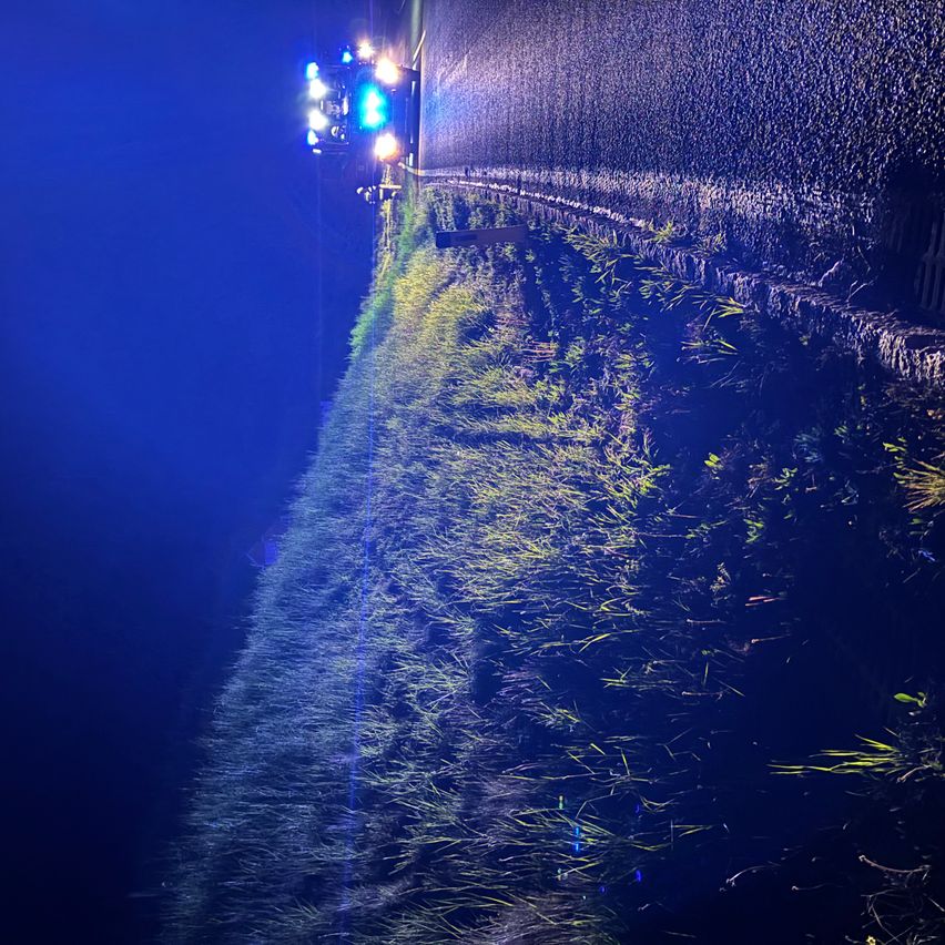 Ein Polizeiauto steht mit eingeschaltetem Blaulicht am Straßenrand. Die Straße wird von Gras und einer Steinmauer gesäumt.