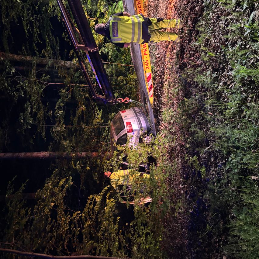 Ein Auto hängt von einem Kran an der Seite einer Straße, wobei zwei Personen in gelben Jacken in der Nähe stehen.