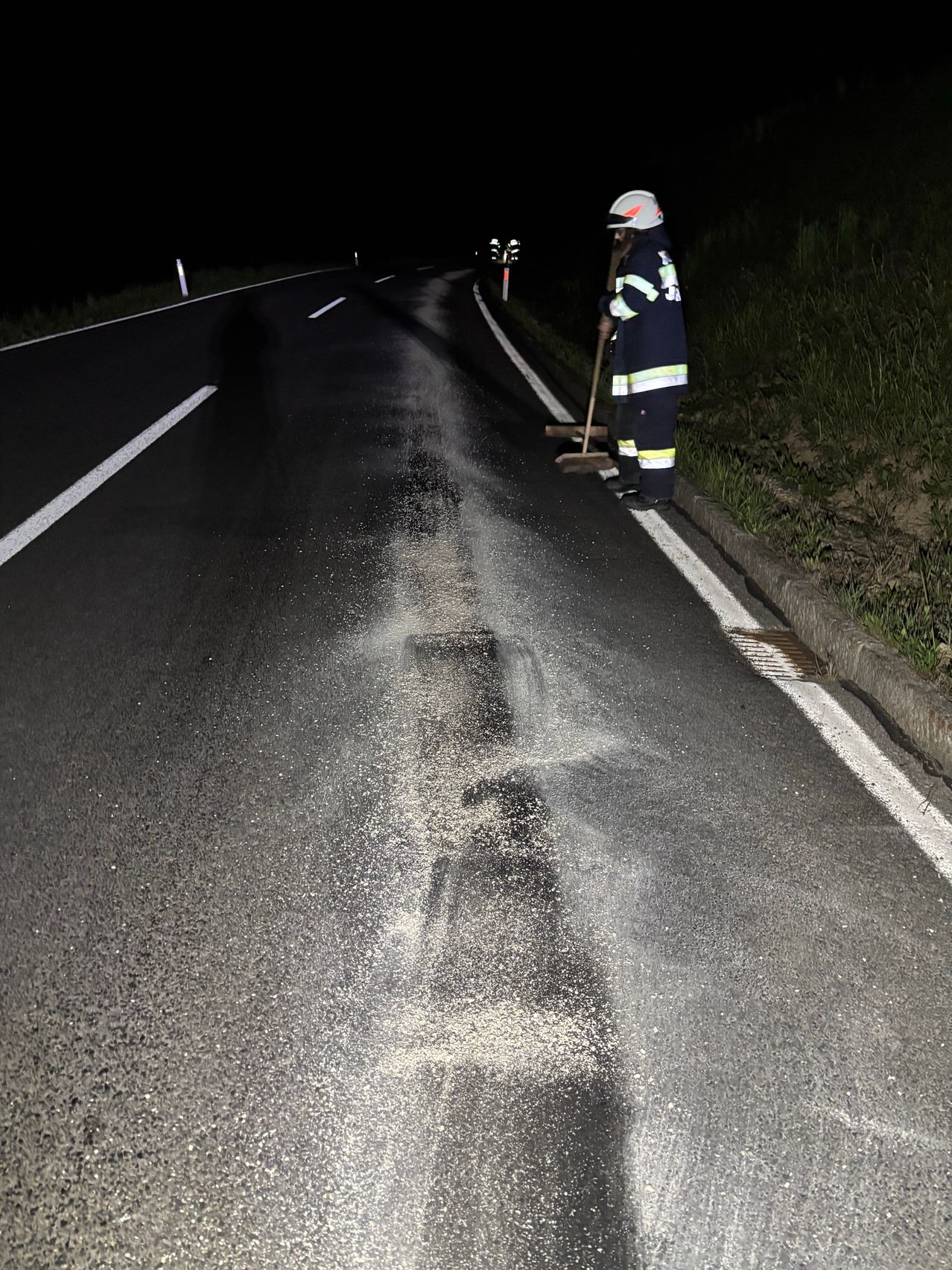 In der Nacht fegt eine Person in Reflektorweste und Helm Schmutz von der Straße. Die Straße hat weiße Markierungen, und der Boden ist nass.