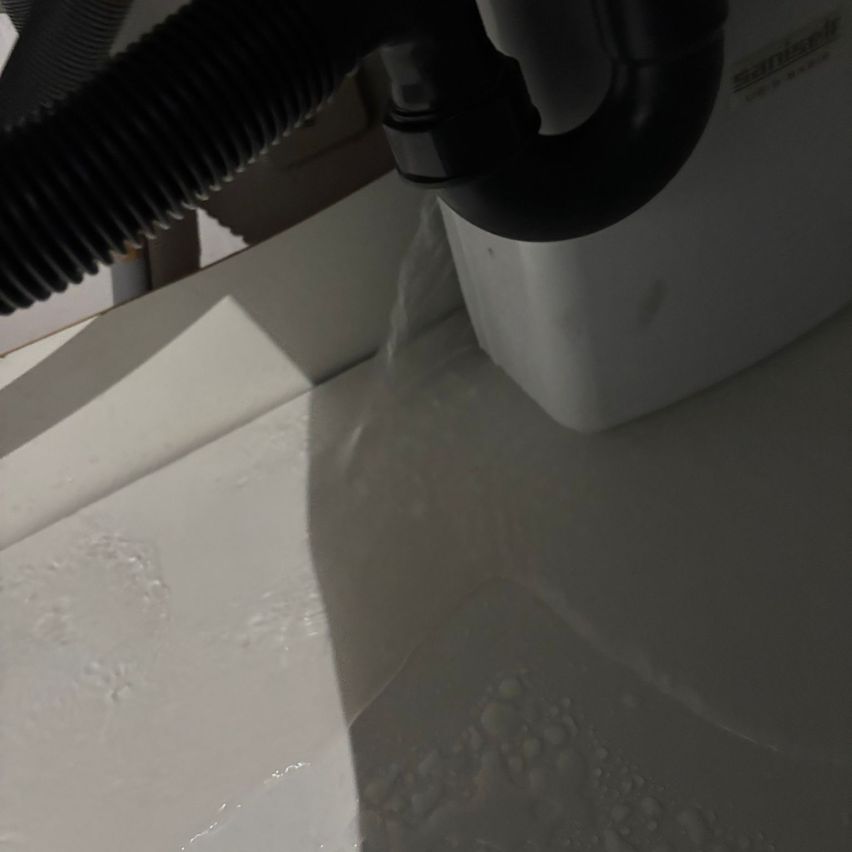 Ein schwarzer Staubsaugschlauch, der mit einem weißen Behälter mit Auslauf verbunden ist und Wasser auf eine weiße Oberfläche verschüttet.