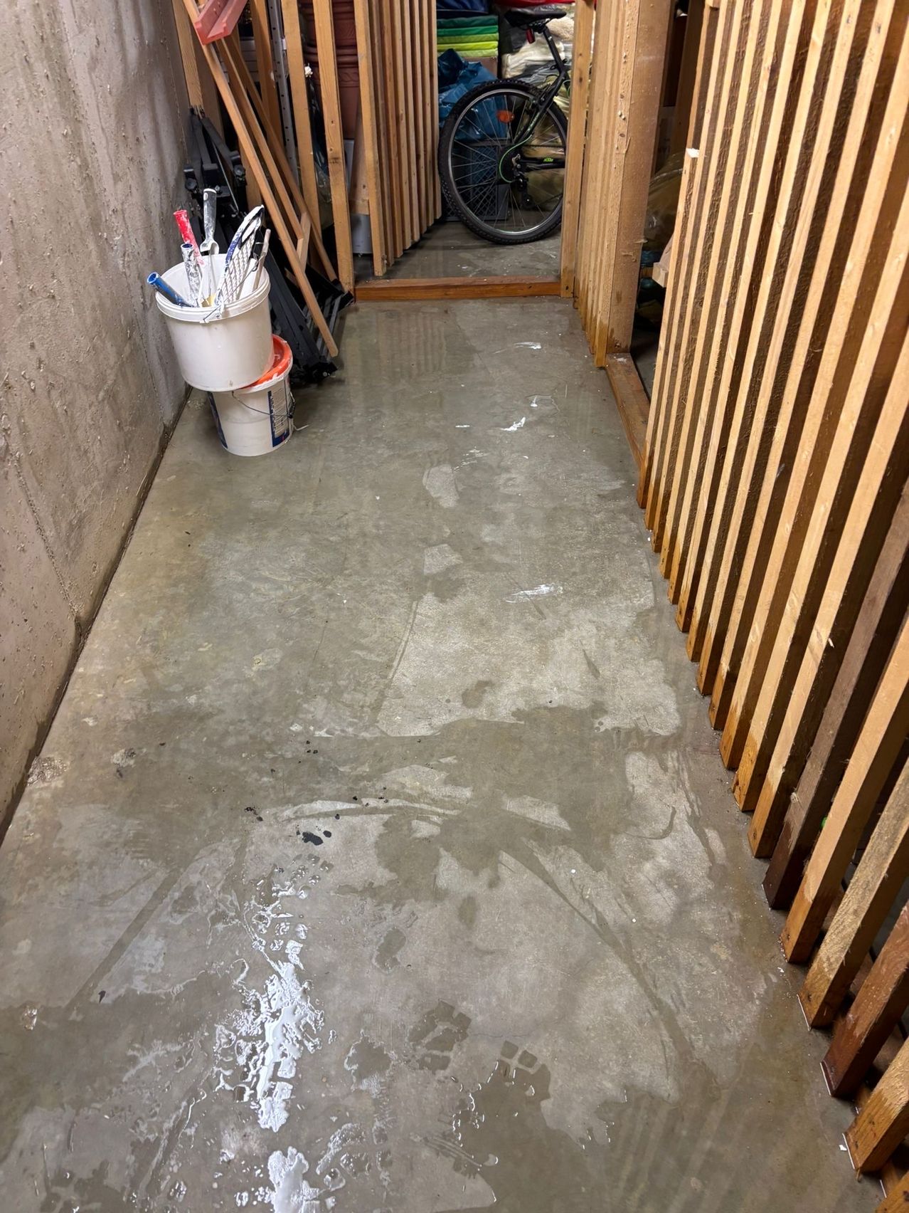 Ein nasser Betonboden in einem Keller mit Eimern und Werkzeugen, die gegen eine Betonwand gestapelt sind. Rechts steht ein Holztor.