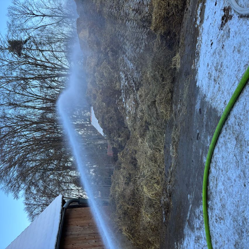 Ein Wassersprüher sprüht Wasser auf einen Heuhaufen. Der Heuhaufen ist von Schnee umgeben, und im Hintergrund sind Bäume zu sehen.