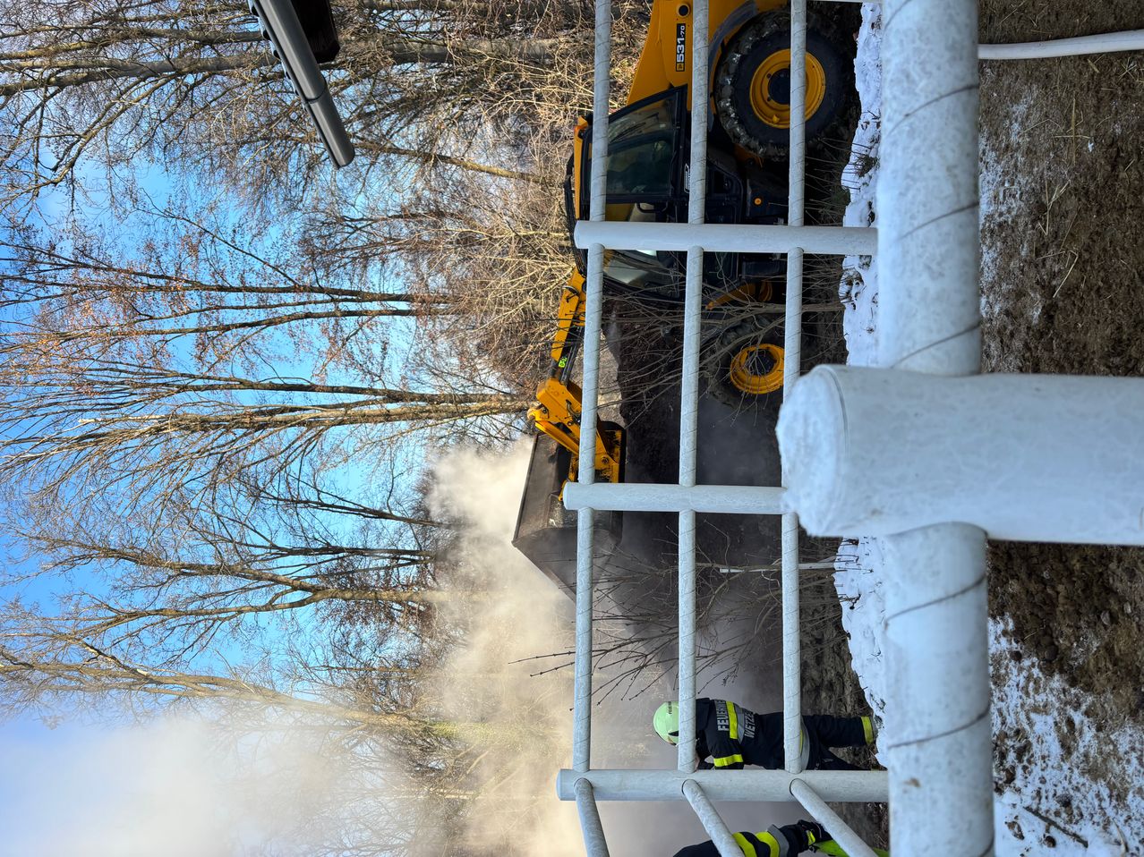 Ein gelber Bagger steht auf einem verschneiten Hang, zwei Arbeiter sind in der Nähe, einer trägt eine gelbe Weste und einen Helm. Der Himmel ist klar und blau.