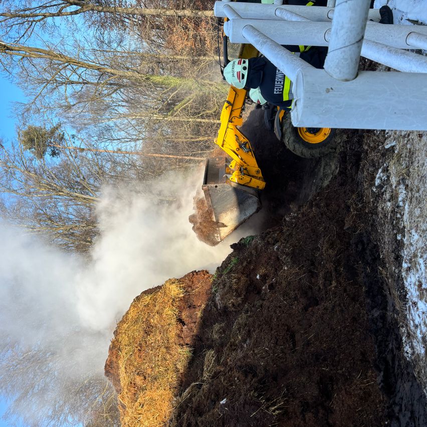 Ein Feuerwehrmann bedient einen gelben Bagger mit einer Schutzbarriere und gräbt in den Boden, wobei Rauch aufsteigt, unter einem klaren blauen Himmel.