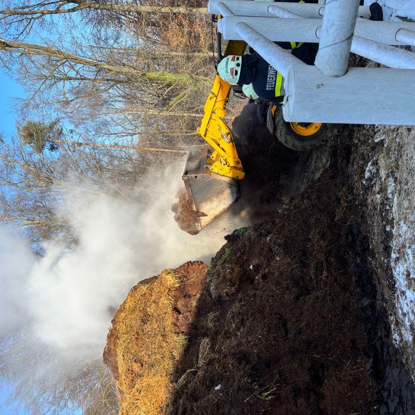 Ein gelber Baumaschinenzug entfernt Erde von einem Haufen. Er stößt Staub aus. Das Gebiet ist von Bäumen umgeben. Der Bediener trägt einen Helm und Schutzkleidung.