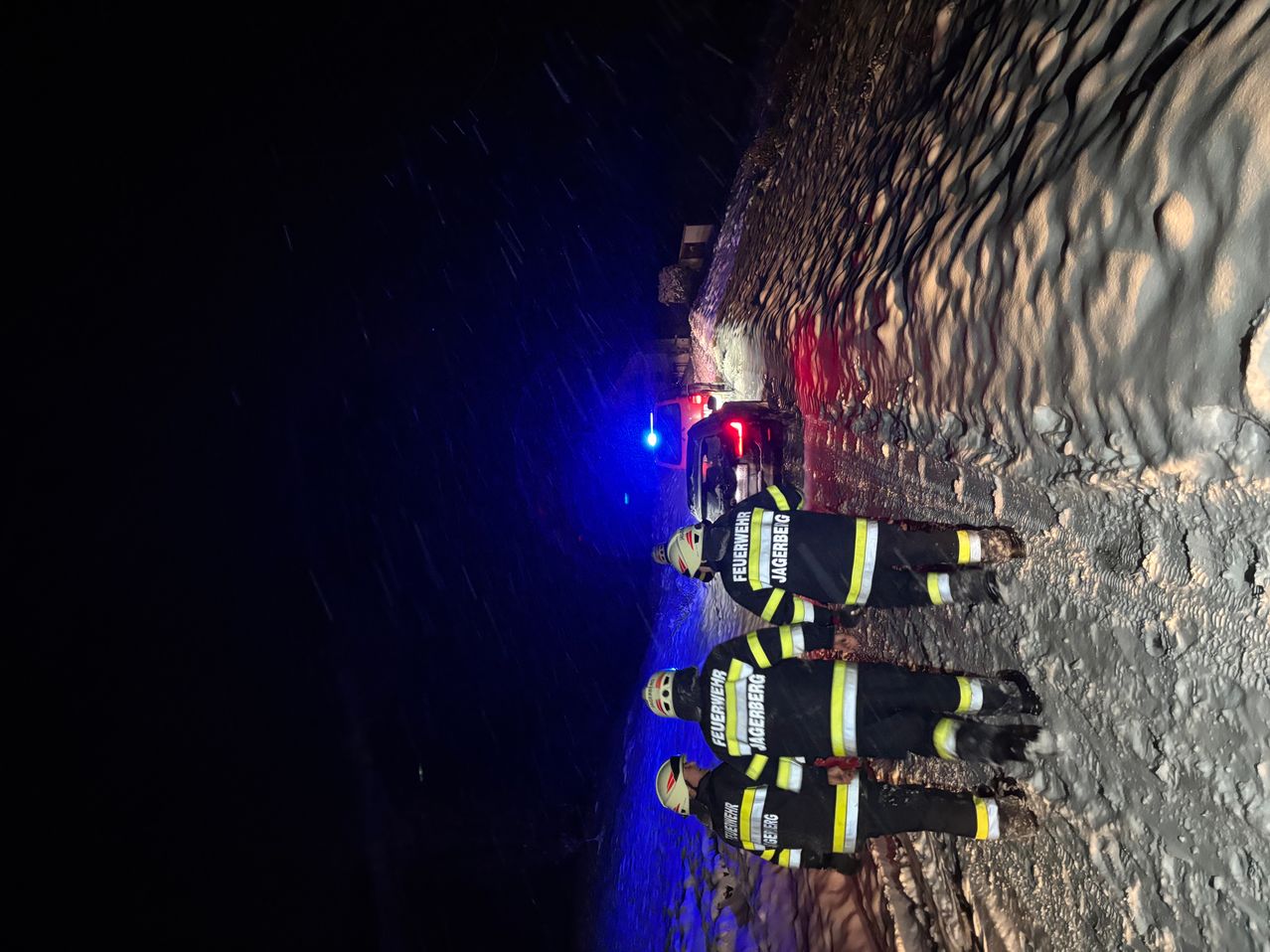 Drei Feuerwehrleute in voller Ausrüstung stehen bei Nacht auf einem verschneiten Hang, wobei ein rotes und blaues Notfalllicht sichtbar ist. Dahinter ist ein Auto geparkt.