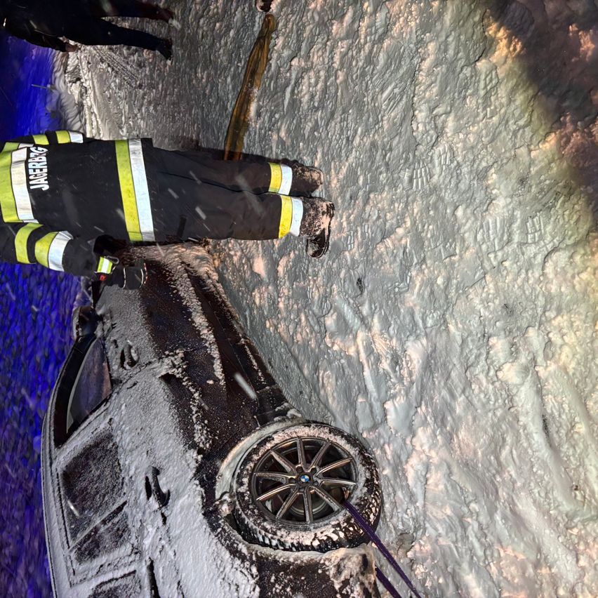 Ein Feuerwehrmann, gekennzeichnet mit JAGERB9, steht neben einem verunglückten Auto, das mit Schnee bedeckt ist, wobei ein Rad abgefallen ist.