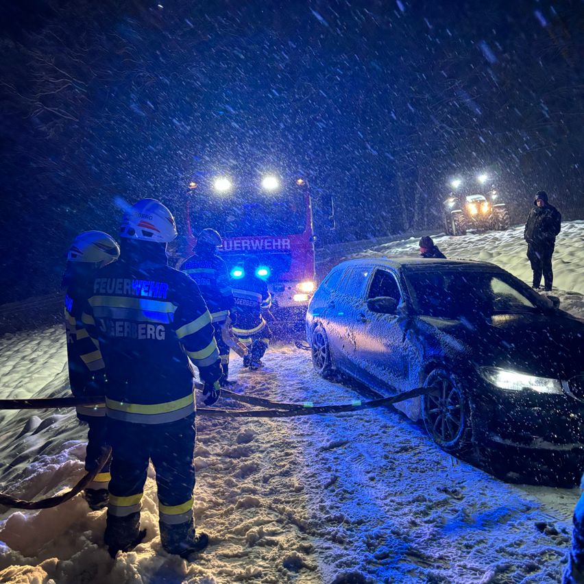 Feuerwehrleute in Schutzkleidung ziehen ein schneebedecktes Auto mit Seilen in einer verschneiten Umgebung. Ein Feuerwehrwagen mit blinkenden Lichtern ist in der Nähe geparkt.