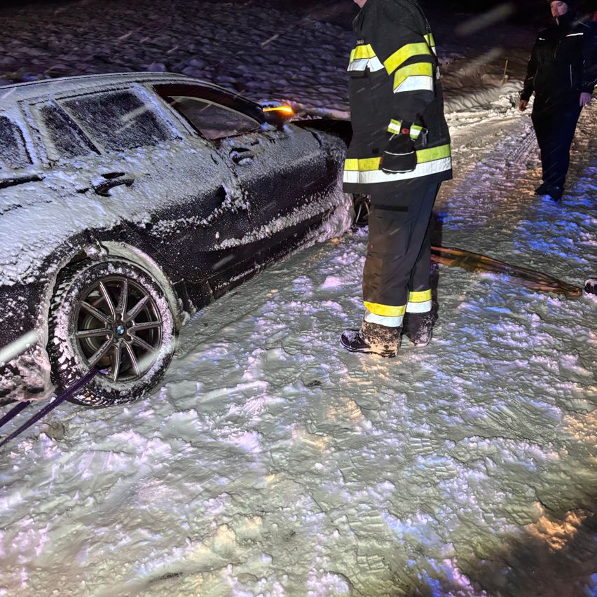 Ein Feuerwehrmann steht neben einem schneebedeckten Auto auf einer verschneiten Straße bei Nacht, mit einer weiteren Person im Hintergrund.