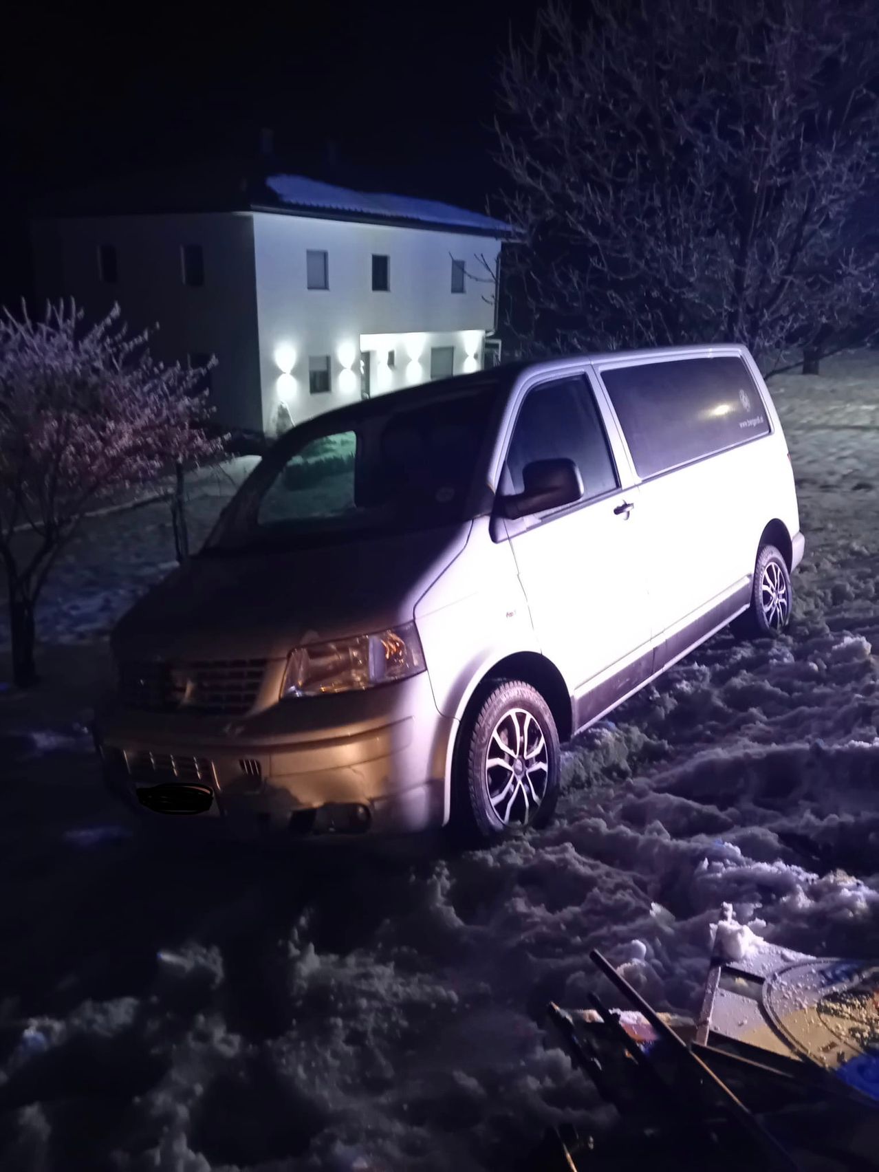 A silver van is parked in the snow at night in front of a house with several windows and lit-up lamps.