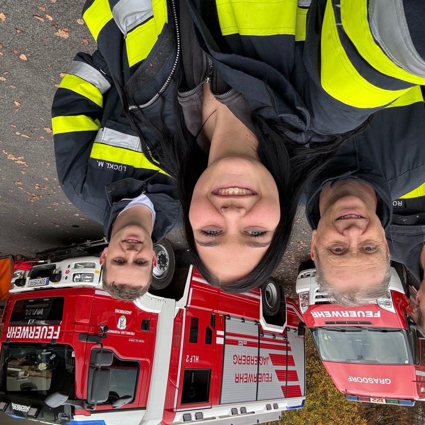 Drei Feuerwehrleute lächeln vor einem Feuerwehrwagen. Die Frau steht in der Mitte, die Männer auf beiden Seiten. Das Fahrzeug ist rot mit weißen Streifen und hat das Wort "Feuerwehr" darauf.