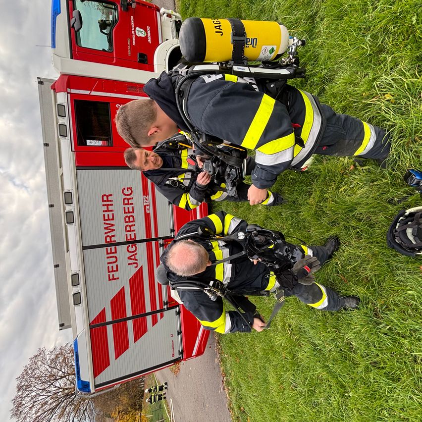 Drei Feuerwehrleute bereiten sich auf einen Einsatz vor, mit Feuerwehrfahrzeugen und Ausrüstung im Hintergrund.