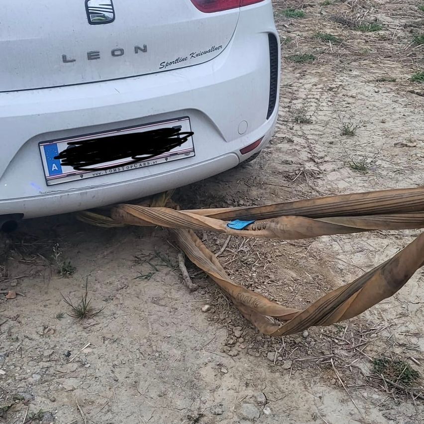 A white Leon car is parked on a muddy road with a license plate obscured by a black mark. A brown rope is tied to the bumper.