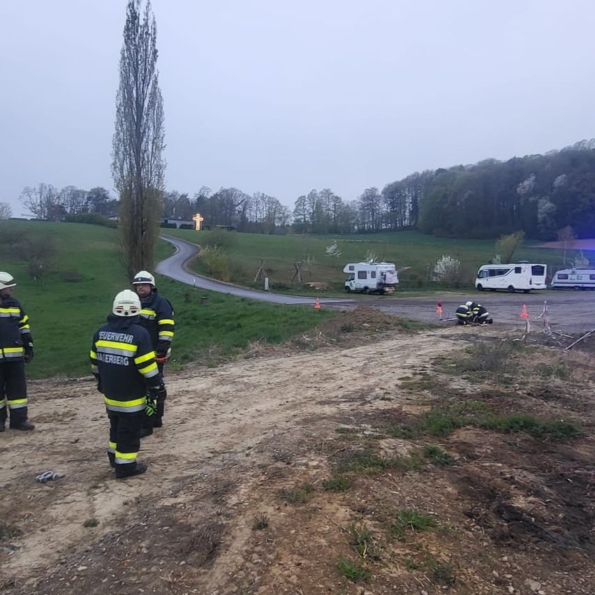 Four firefighters stand on a dirt path near a grassy field. In the background, RVs and emergency vehicles are parked.