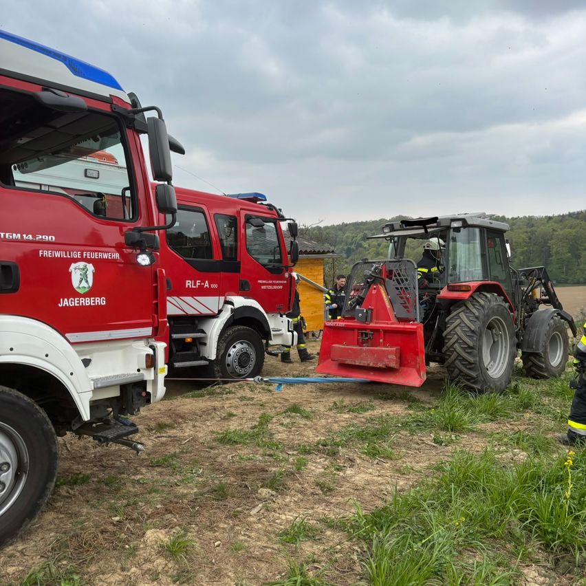 Fire trucks and a tractor are parked on a grassy field under a cloudy sky. A person sits on the tractor, and others are near the fire truck.