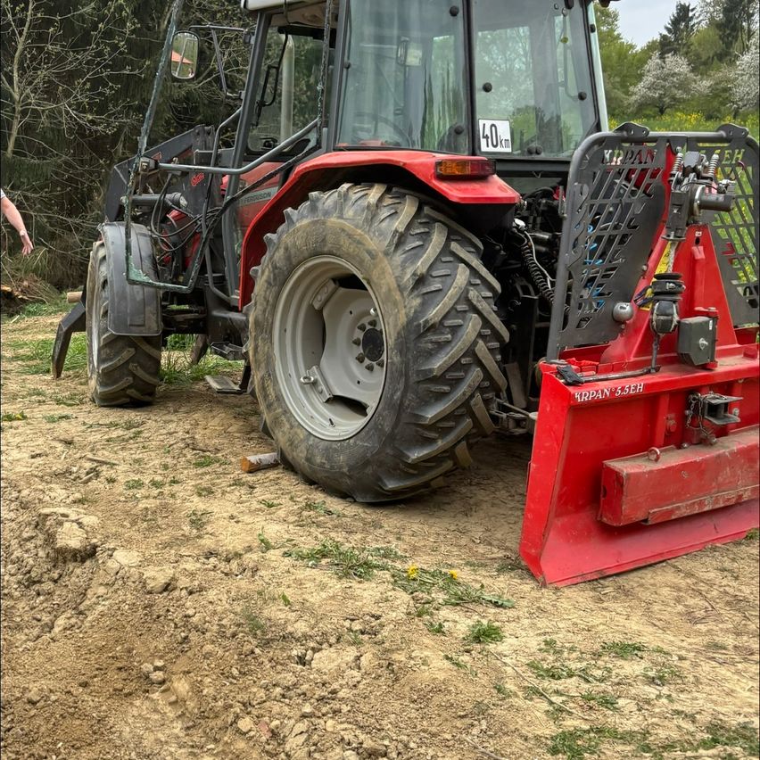 A red tractor with a 40km plate is parked on a dirt field with trees and a person nearby.