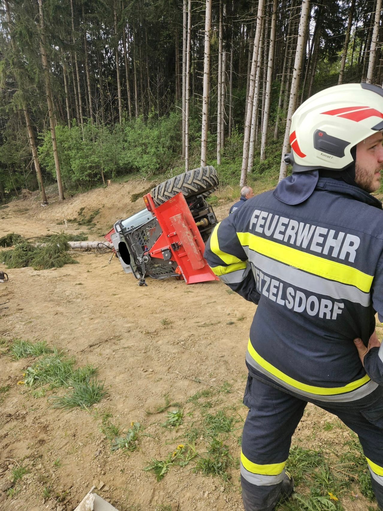 A firefighter from Mittelsdorf is standing next to an overturned vehicle. A person is nearby. The area is surrounded by trees.