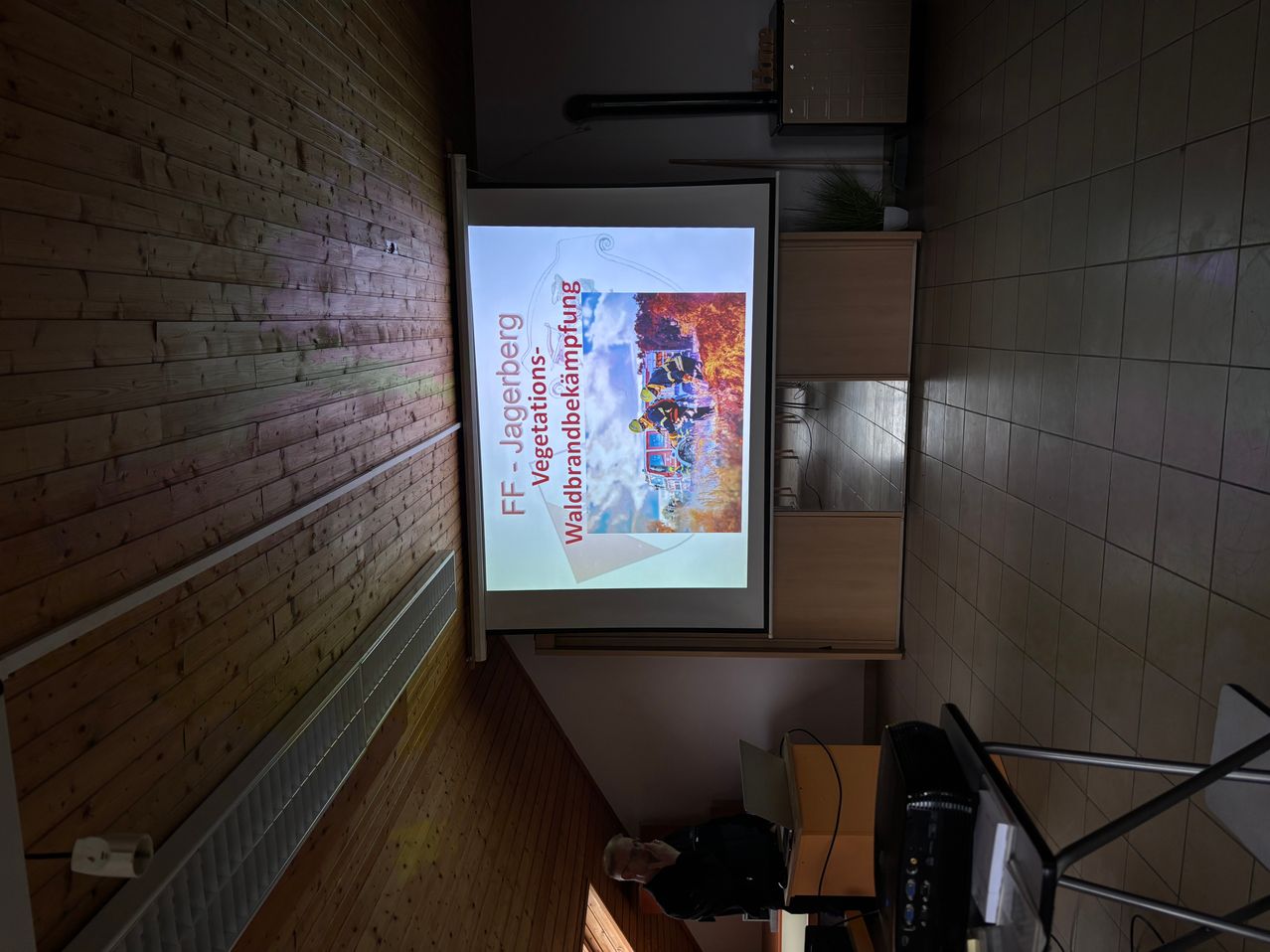 Ein Raum für Konferenzen hat eine Projektionswand, die eine Präsentation über Feuerwehrleute zeigt. Ein Mensch sitzt in der Nähe mit einem Laptop.