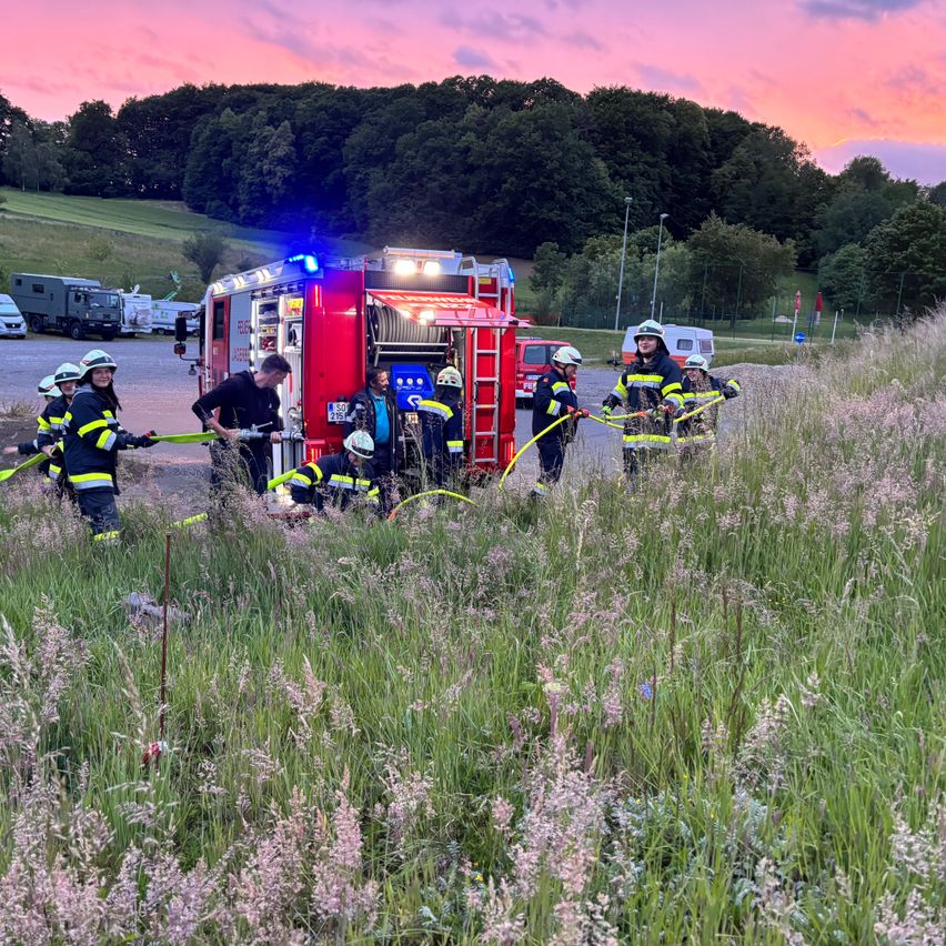 Eine Gruppe von Feuerwehrleuten in Schutzausrüstung arbeitet an einem Feuerwehrauto und spüht Wasser in hohes Gras. Im Hintergrund befinden sich Fahrzeuge und ein Gebäude.