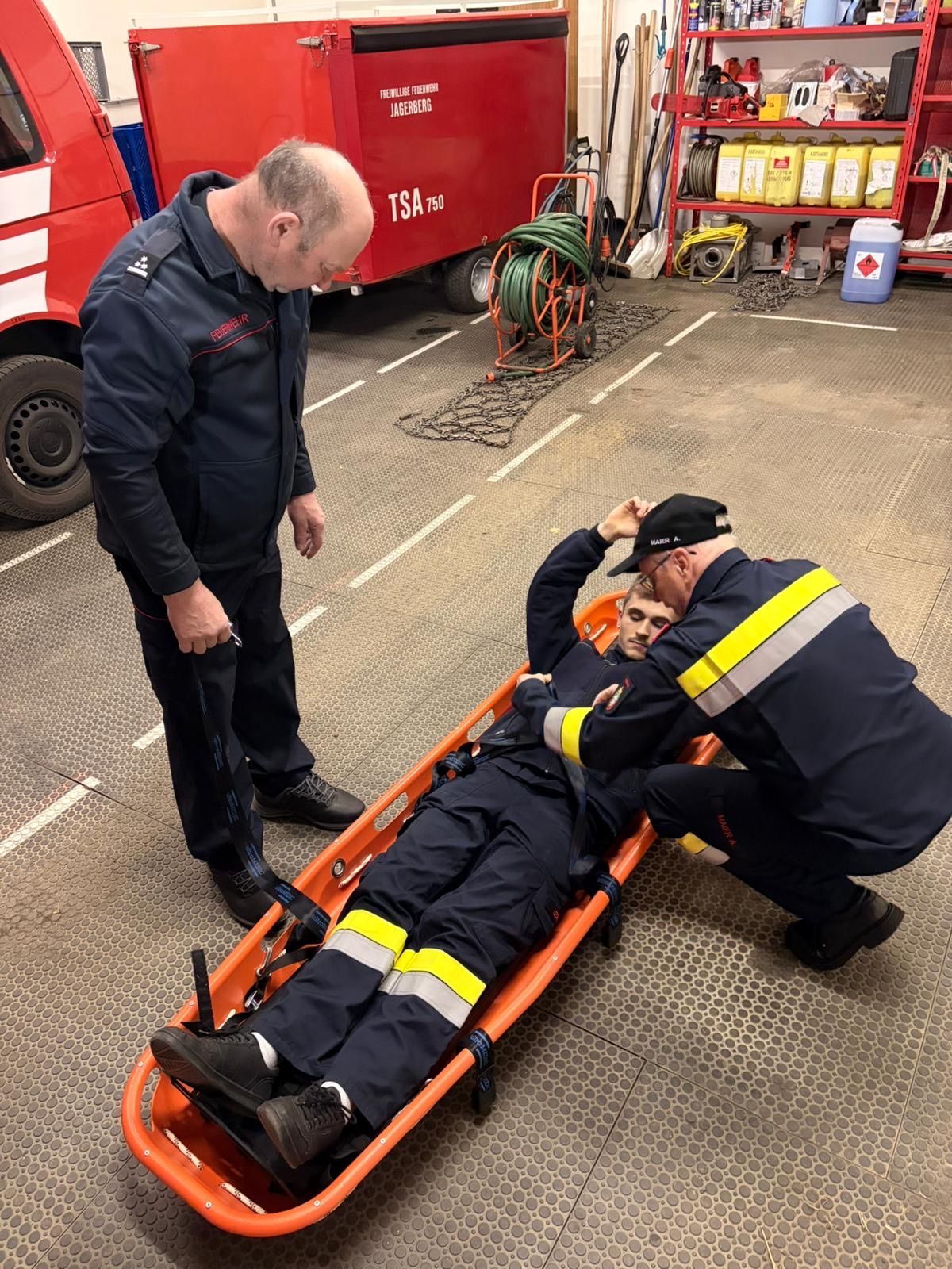 Zwei Feuerwehrleute sichern einen Mann auf einer Trage in einer Garage. Ein Feuerwehrmann schaut den Mann an und der andere steht und hält ein Werkzeug.