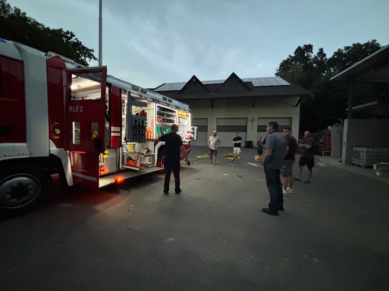 A fire truck with its door open, labeled HLF 2, parked in front of a building with solar panels. Several people are standing around, some near the truck and others near the building.