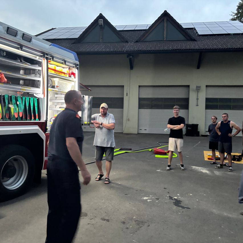 A group of men stand in a parking lot, some wearing hats and sneakers, with a fire truck and a building with solar panels in the background.