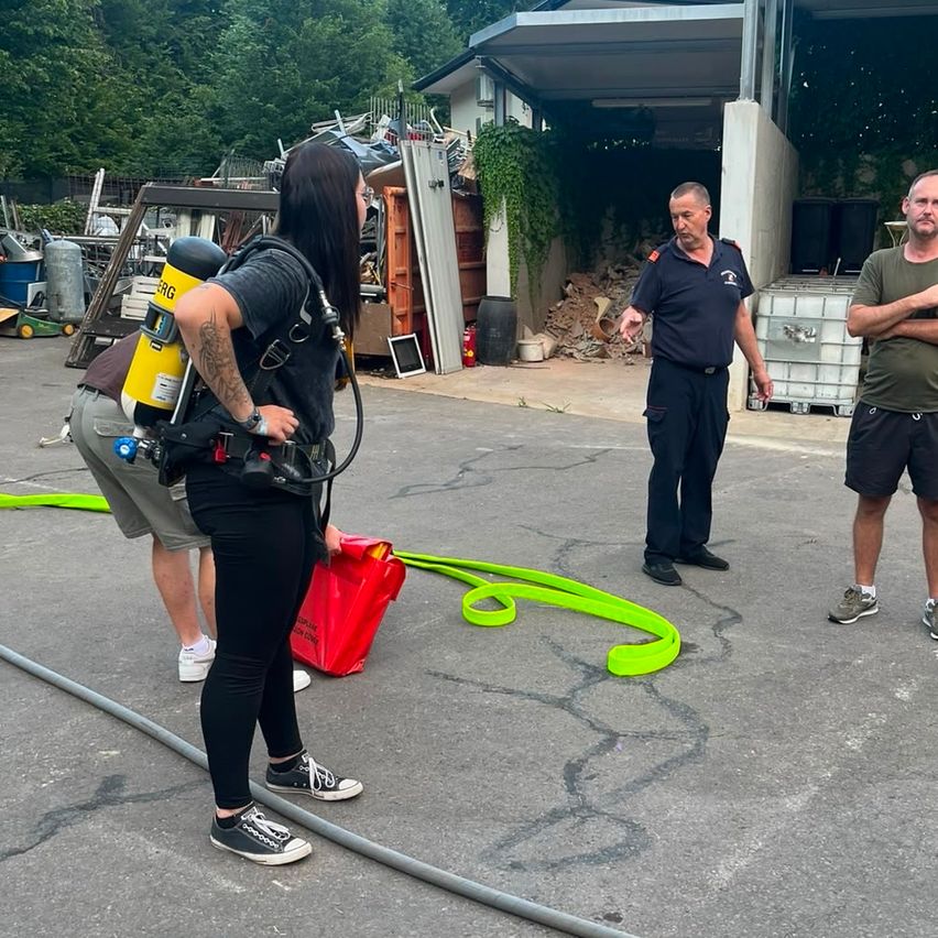 Three individuals are outside a building with a garage. A woman is wearing a diving suit, and two men are standing with their arms crossed. A red bag and a green hose are on the ground.