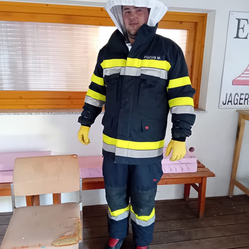 Ein Mann in einer reflektierenden Schutzkleidung steht in einem Raum mit Holzboden und Fenster. Er trägt gelbe Handschuhe und eine Kapuze.