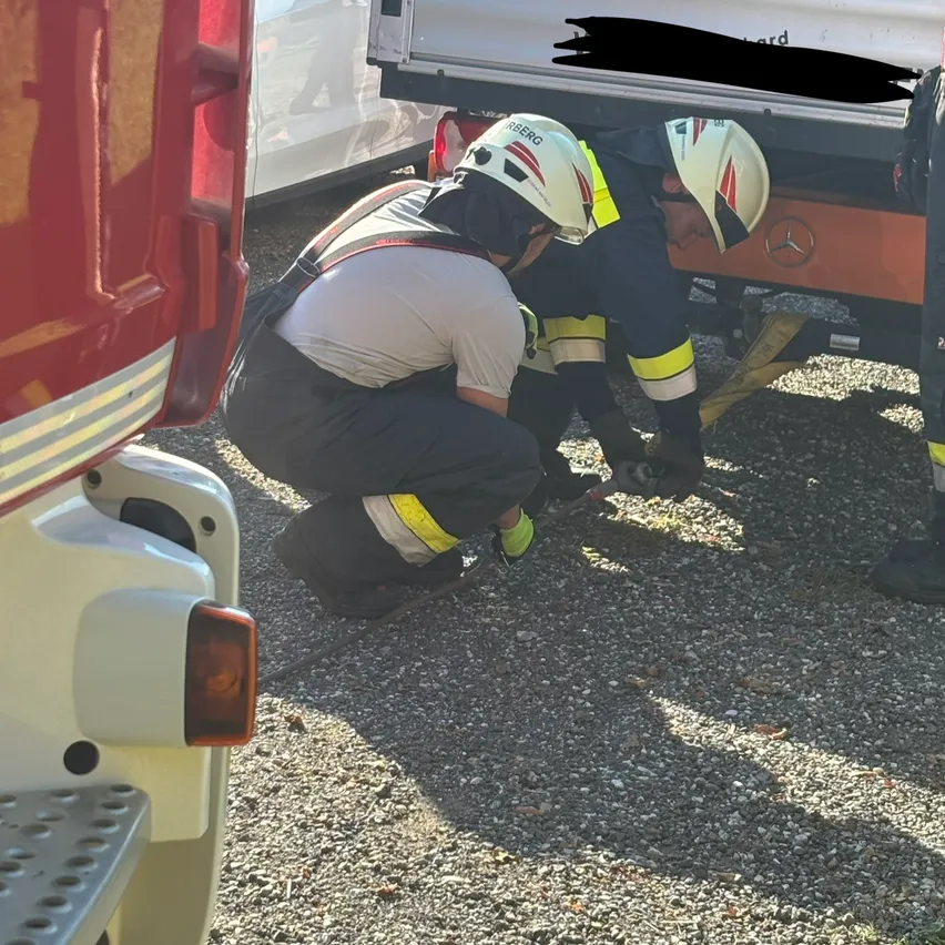 Zwei Feuerwehrleute in voller Ausrüstung arbeiten am Heck eines Feuerwehrwagens, einer kniet und der andere beugt sich vor. Die Szene spielt sich im Freien auf einer Kiesoberfläche ab.