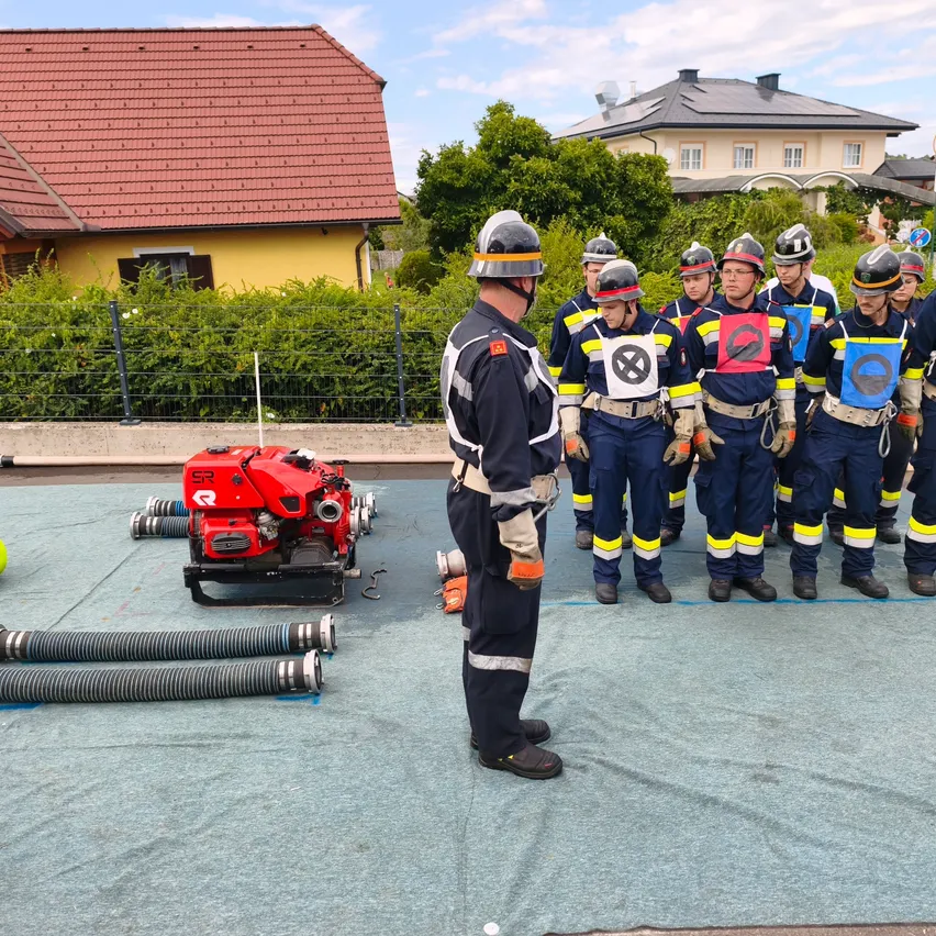 Eine Gruppe von Feuerwehrleuten in Uniform steht in einer Reihe, trägt Helme und hält Ausrüstung. Ein Mann vor ihnen scheint Anweisungen zu geben. Hinter ihnen sind Häuser und ein rotes Maschinenfahrzeug.