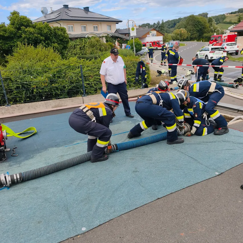 Feuerwehrleute arbeiten auf einer Straße, einer benutzt einen Schlauch, während andere helfen. Ein Vorgesetzter steht in der Nähe, und ein Haus und Fahrzeuge sind im Hintergrund.