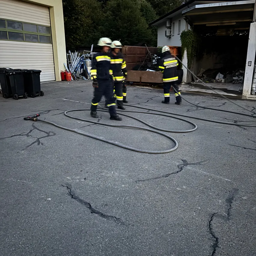 Drei Feuerwehrleute in voller Ausrüstung gehen auf einer rissigen Straße und halten Schläuche. Sie sind in der Nähe eines Gebäudes mit offener Garagentür.