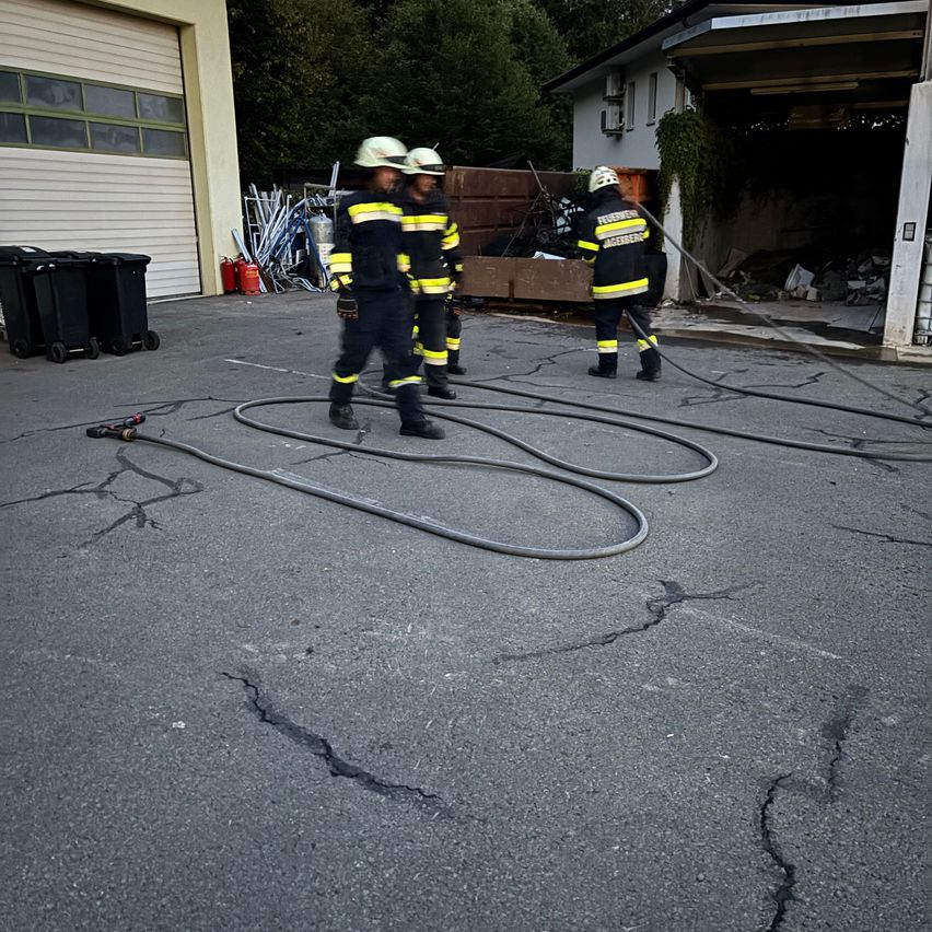 Drei Feuerwehrleute in voller Ausrüstung gehen auf einer rissigen Straße und halten Schläuche. Sie sind in der Nähe eines Gebäudes mit offener Garagentür.
