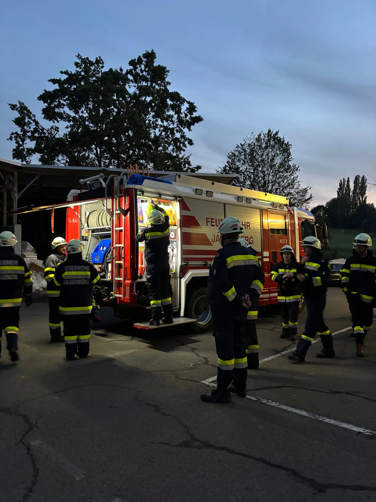 Feuerwehrleute in voller Montur stehen um einen Feuerwehrwagen herum, einer klettert die Leiter hinauf. Die Szene spielt sich auf einem Parkplatz mit Bäumen im Hintergrund ab.