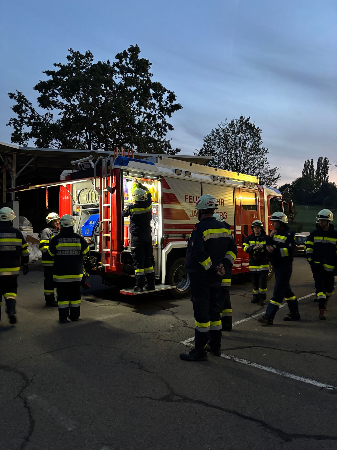 Feuerwehrleute in voller Montur stehen um einen Feuerwehrwagen herum, einer klettert die Leiter hinauf. Die Szene spielt sich auf einem Parkplatz mit Bäumen im Hintergrund ab.