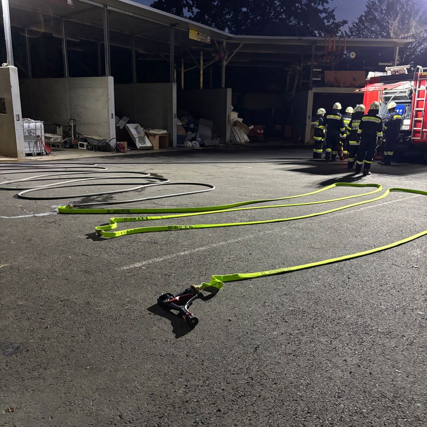 Feuerwehrleute stehen nachts in einer Garage, mit Feuerwehrschläuchen und Ausrüstung um sie herum. Ein roter Feuerwehrwagen ist in der Nähe geparkt.