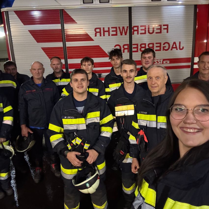Eine Gruppe von Feuerwehrleuten in Uniform, einige halten Helme, posiert für ein Foto vor einem Feuerwehrwagen.