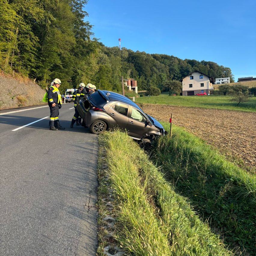 Ein Auto ist am Straßenrand beschädigt und wird von Rettungskräften versorgt. Die Szene spielt sich in einer ländlichen Gegend mit einem Haus im Hintergrund ab.